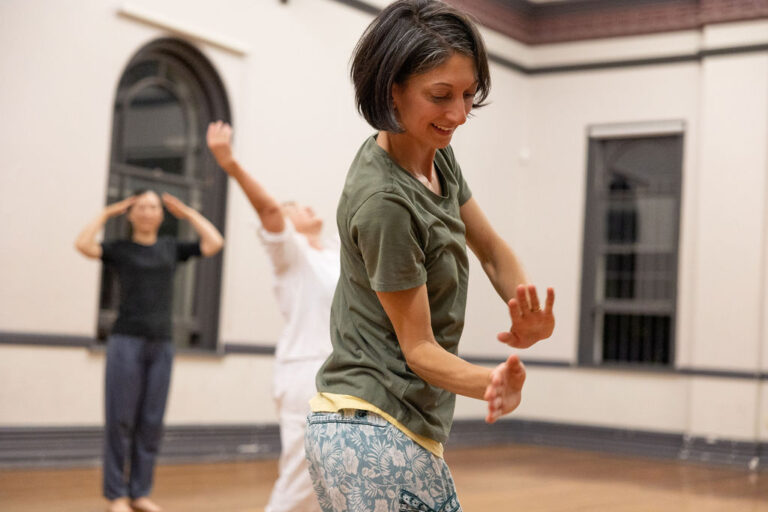 Young women in her 30s with dark brown short hair, wearing a green tshirt and blue pants stand side on to the photo. Her hands with fingers towards the sky near her right hip. Smiling with her eyes looking down towards her hands.