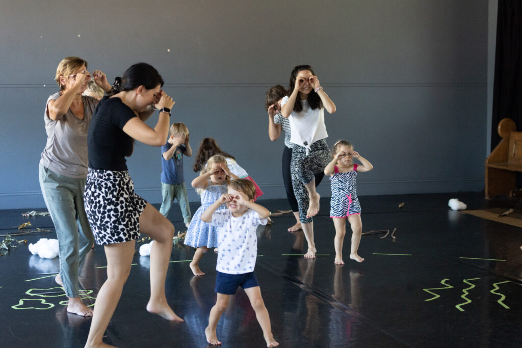 Group of mothers, grandmother and children walking in a dance studio with their hands cupping their eyes as if they are wear binoculars.