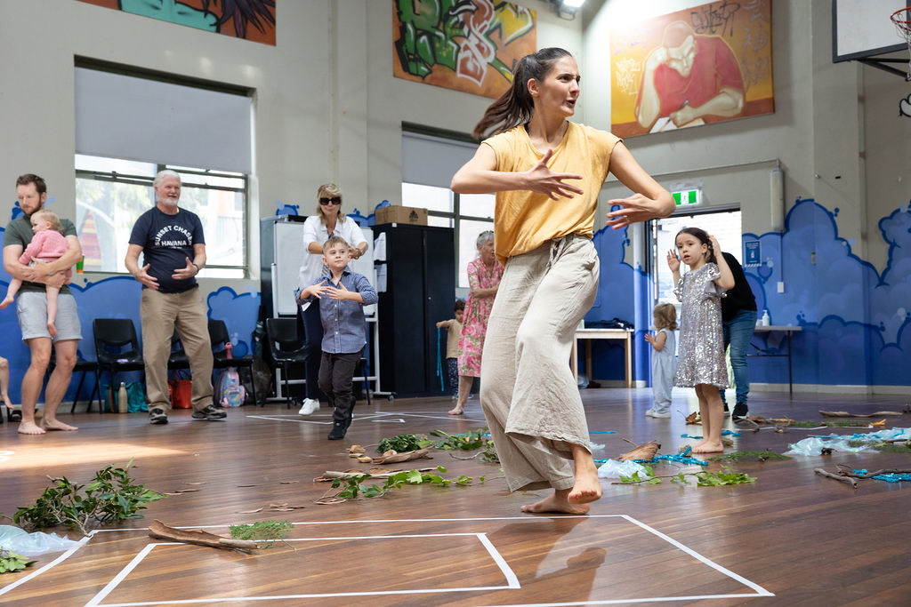 Lead facilitator in yellow and green clothes centre foreground of picture- twisting her body with her arms in front of her. Seniors, Parents and Children seen in background dancing with her. Leaves, sticks and branches with some bright coloured blue ribbon all over the floor.